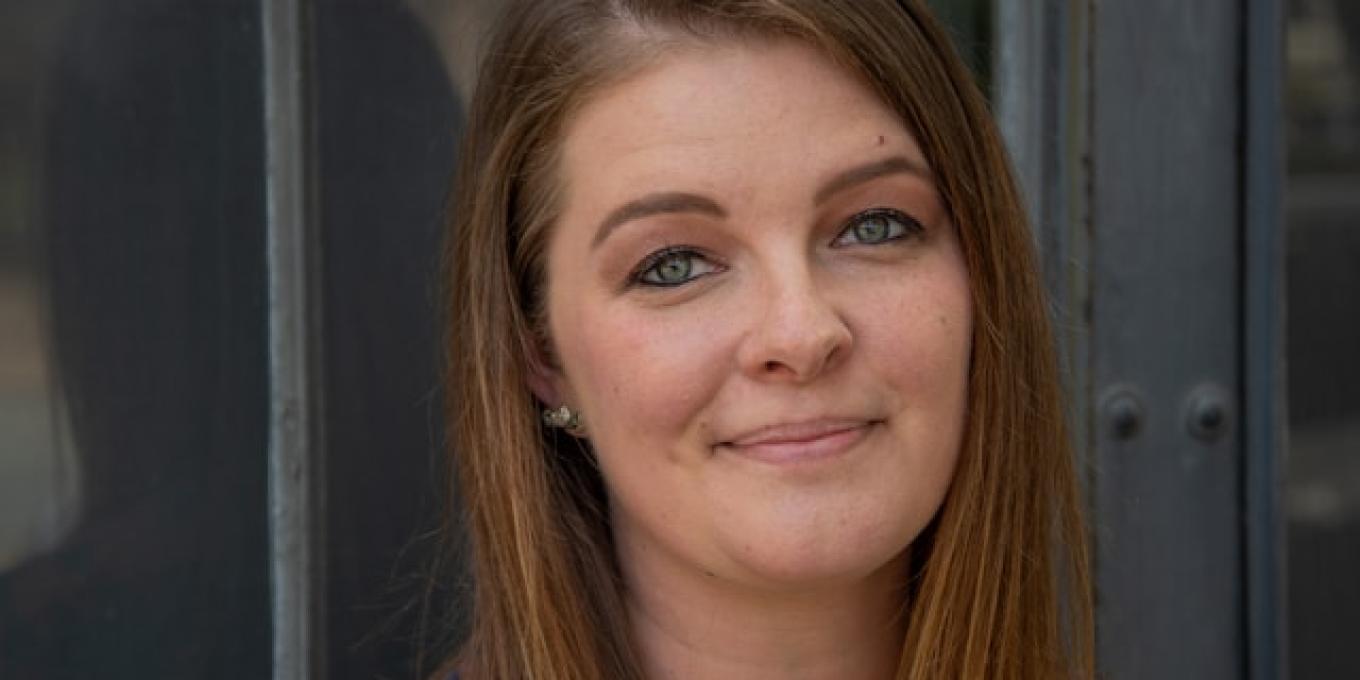 Smiling woman in a patterned shirt, standing by a window.